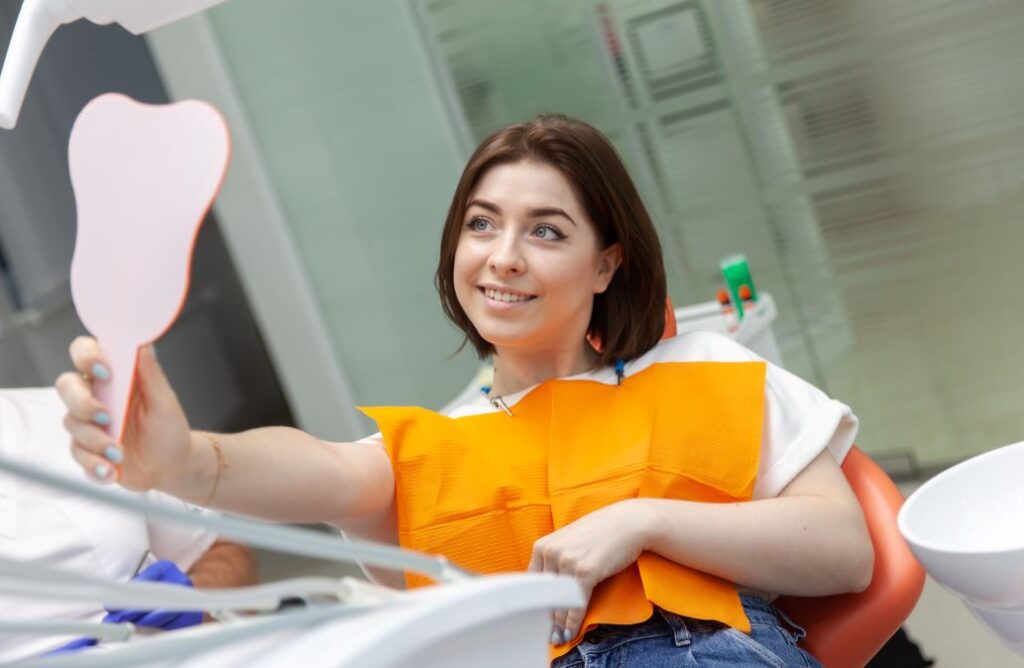 A woman in a dentist’s chair looking at her teeth in a hand mirror.